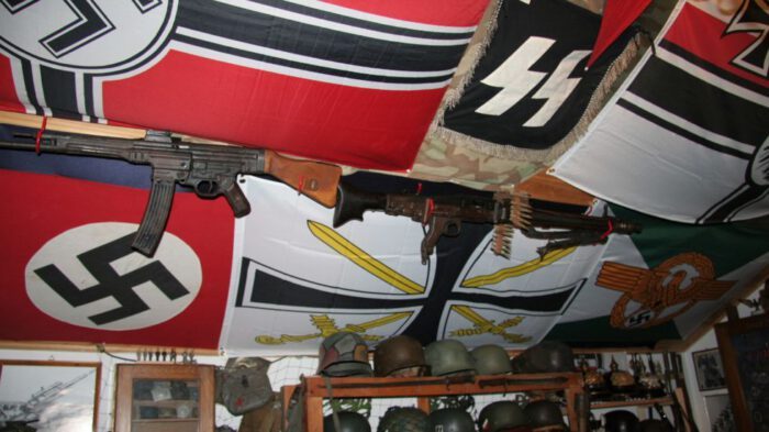 Zwei historische Gewehre hängen unter einer Decke mit verschiedenen Flaggen, darunter NS- und Wehrmachtsymbole.