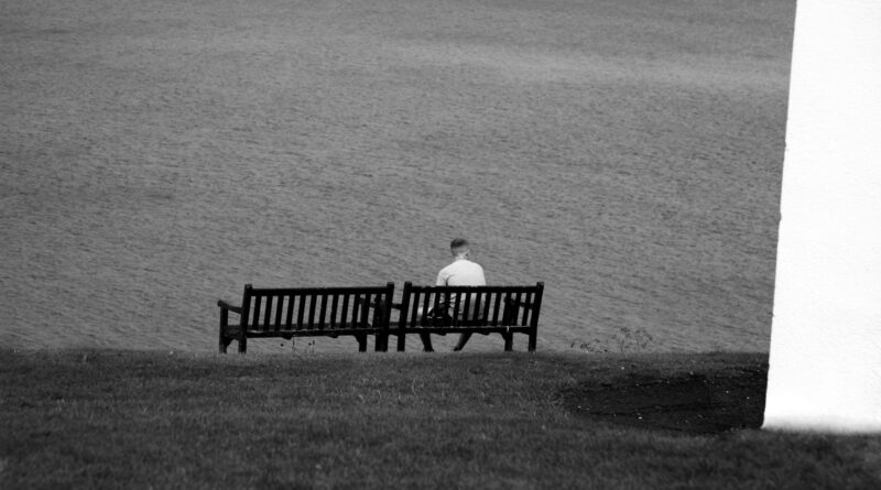 man sitting on bench facing the body of water during day