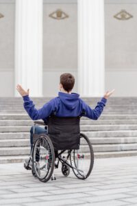 Young man in a wheelchair facing stairs, arms raised, outdoors.