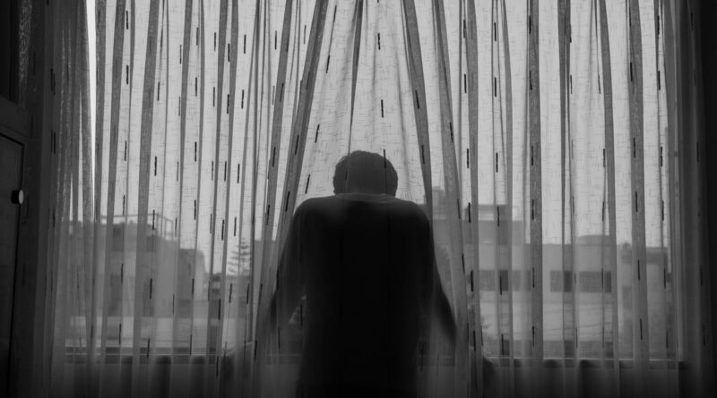 A black and white photo of a person standing behind a curtain, looking out the window, conveying isolation.